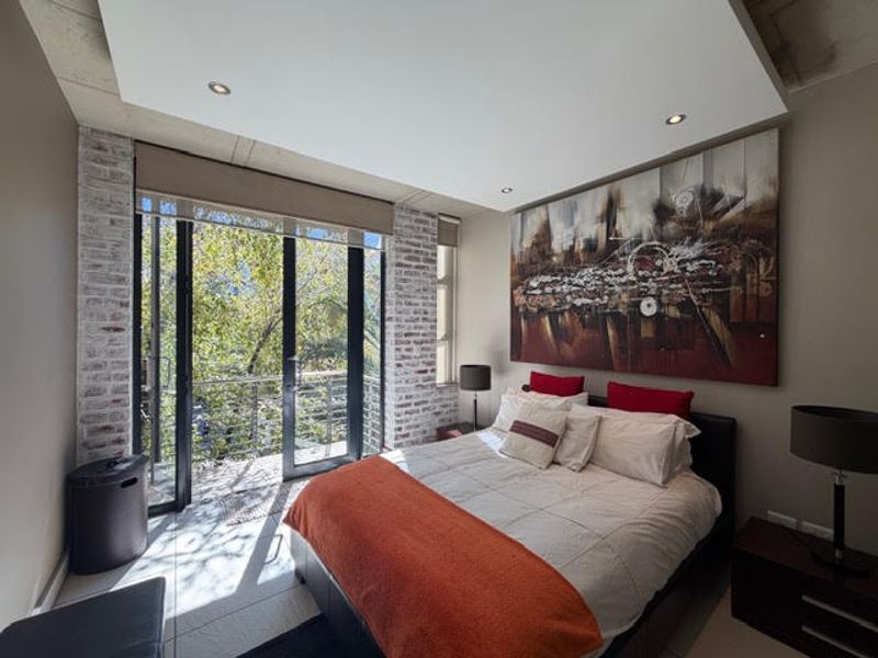 Modern bedroom featuring exposed brick accents, recessed ceiling lighting, and large sliding glass doors opening onto a private balcony with natural views.