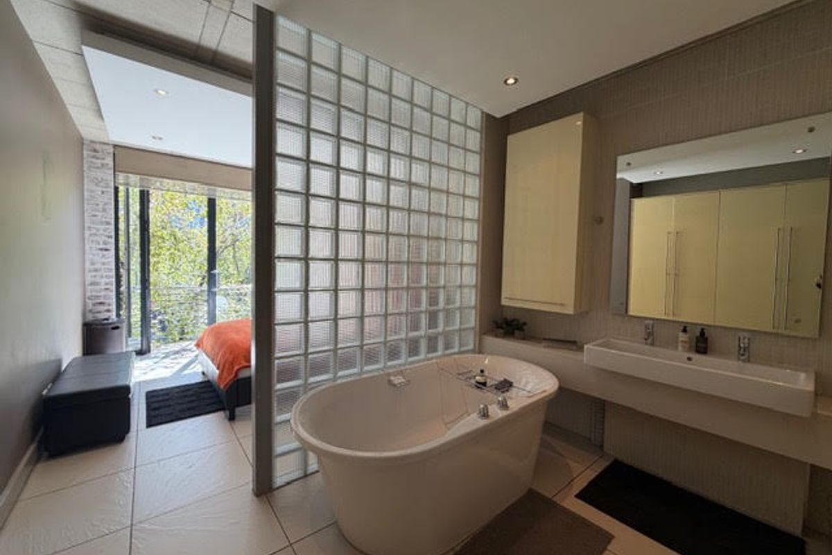 Modern open-plan bathroom featuring a freestanding bathtub, glass block partition, double vanity, and direct access to a balcony area.