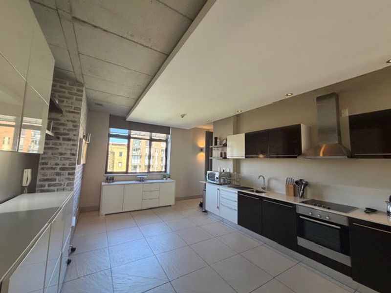 Modern open-plan kitchen featuring sleek cabinetry, exposed concrete ceilings, a brick feature wall, and high-quality built-in appliances in a contemporary apartment setting.