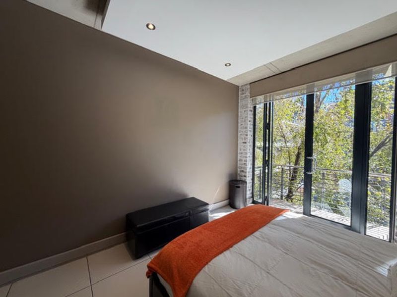 Modern bedroom featuring exposed concrete ceilings, recessed lighting, an exposed brick accent column, and large glass sliding doors opening to a balcony.