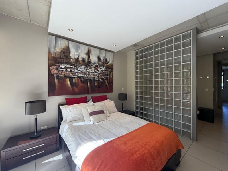 Modern bedroom featuring a distinctive glass block partition wall, recessed downlighting, and neutral tiled flooring, creating a bright and contemporary living space.