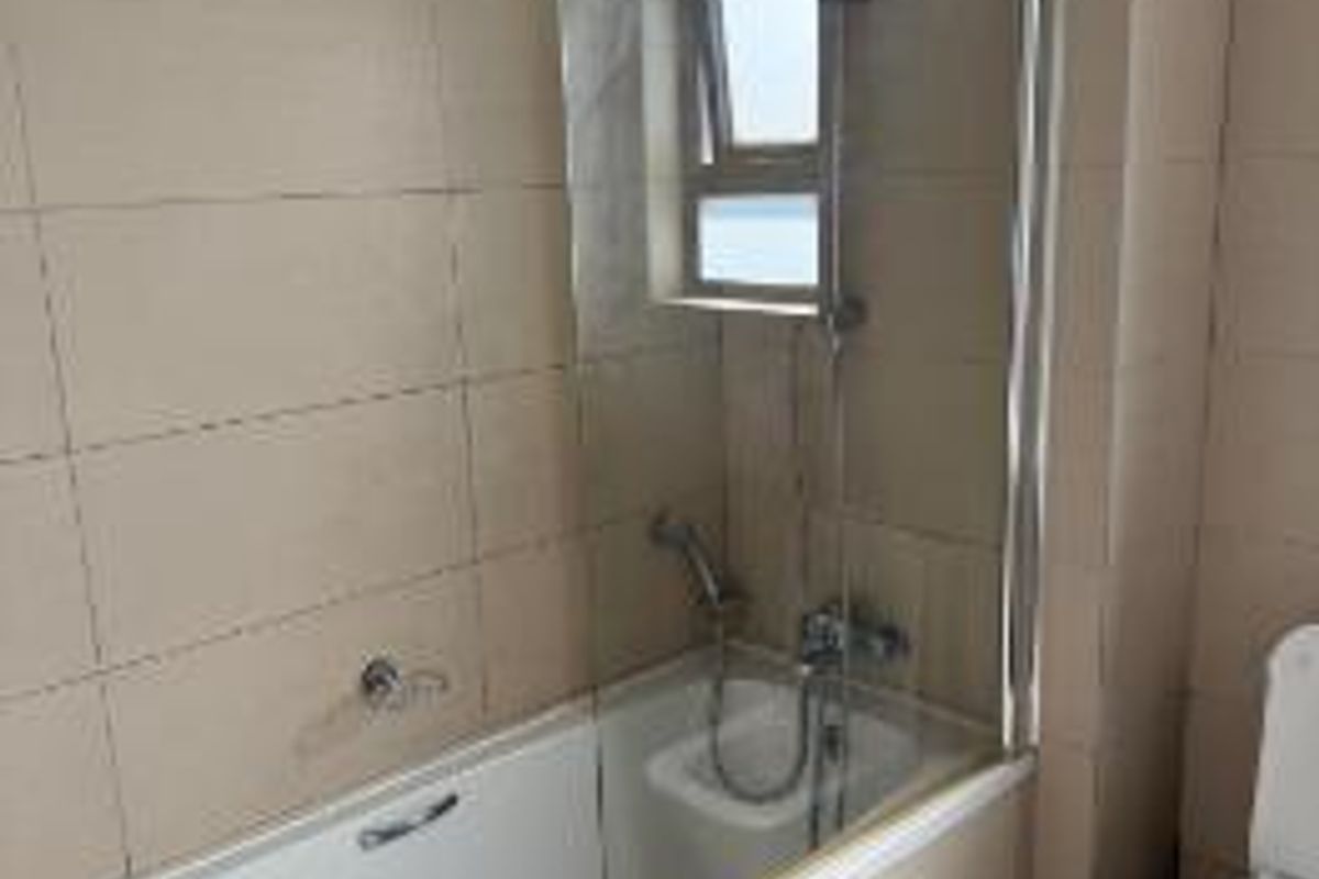 A functional bathroom featuring a bathtub with a glass shower screen, handheld shower head, and a standard toilet with neutral wall and floor tiling.