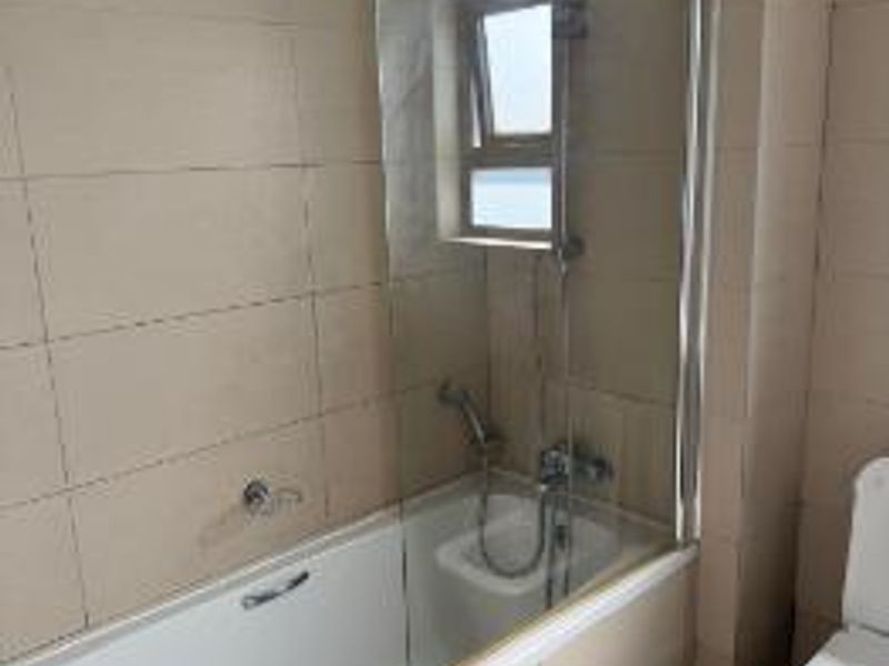 A functional bathroom featuring a bathtub with a glass shower screen, handheld shower head, and a standard toilet with neutral wall and floor tiling.
