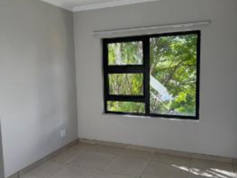 A bright, empty bedroom featuring neutral tiled flooring, clean grey walls, and a modern black aluminium window frame overlooking greenery.