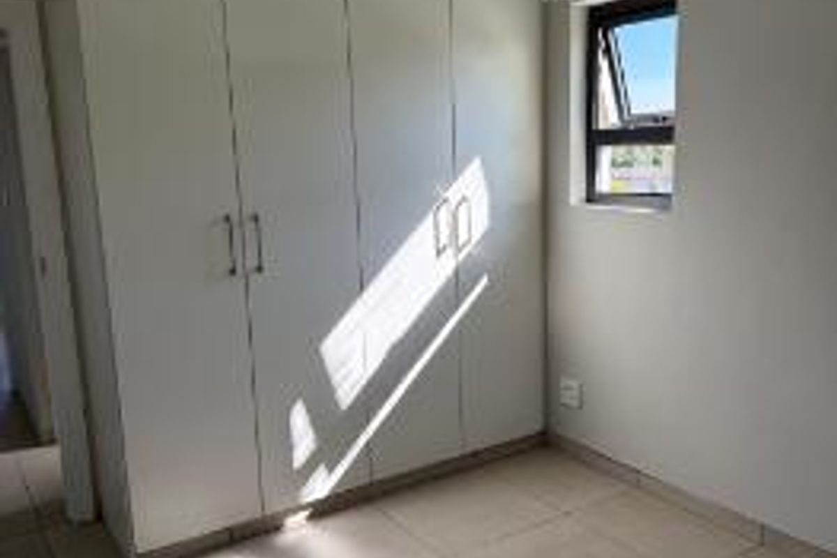 A bright, empty bedroom featuring neutral tiled flooring, built-in white cupboards, and a single window providing natural light.