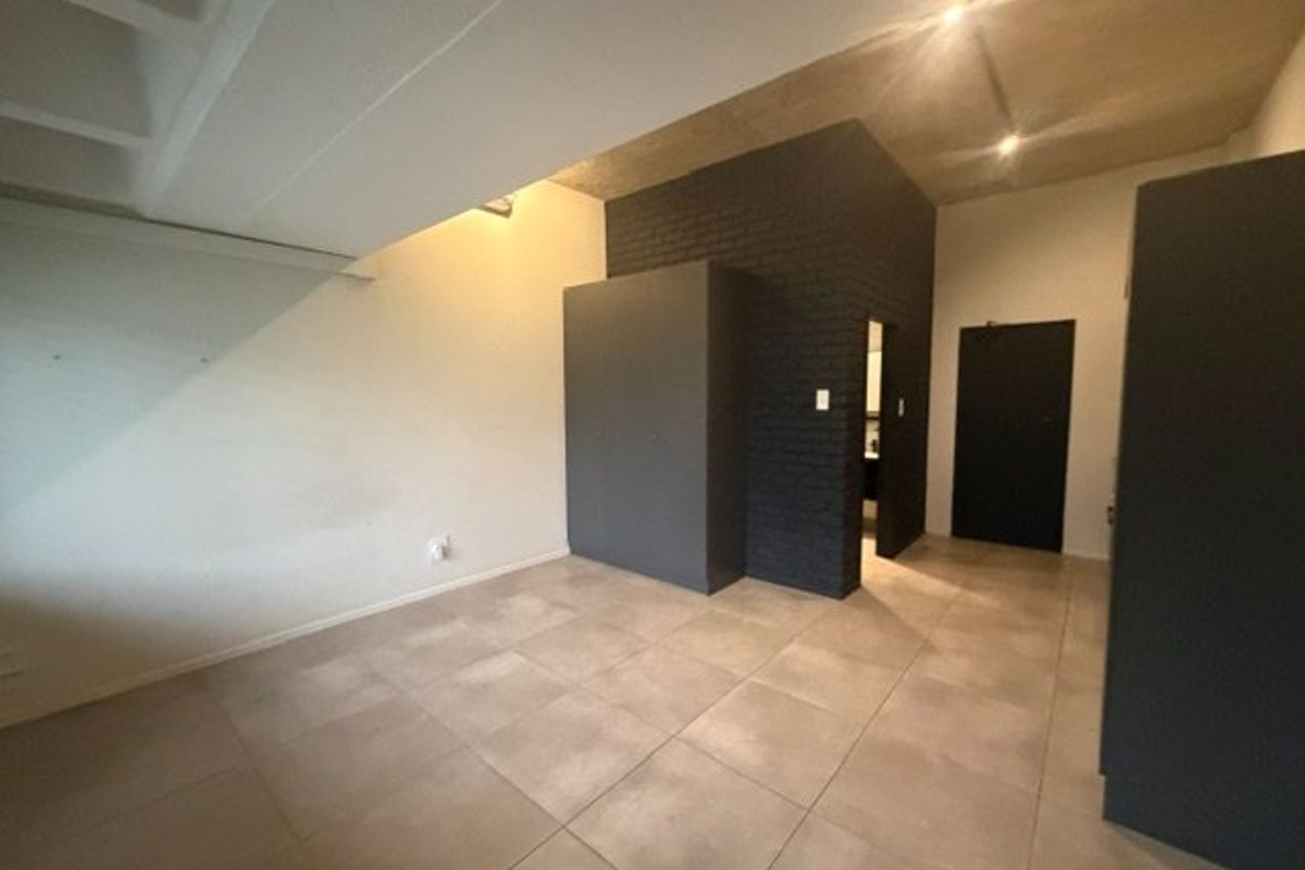 A modern, minimalist bedroom featuring an exposed concrete ceiling, tiled flooring, and a striking dark painted brick feature wall with built-in storage.