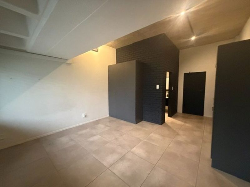 A modern, minimalist bedroom featuring an exposed concrete ceiling, tiled flooring, and a striking dark painted brick feature wall with built-in storage.