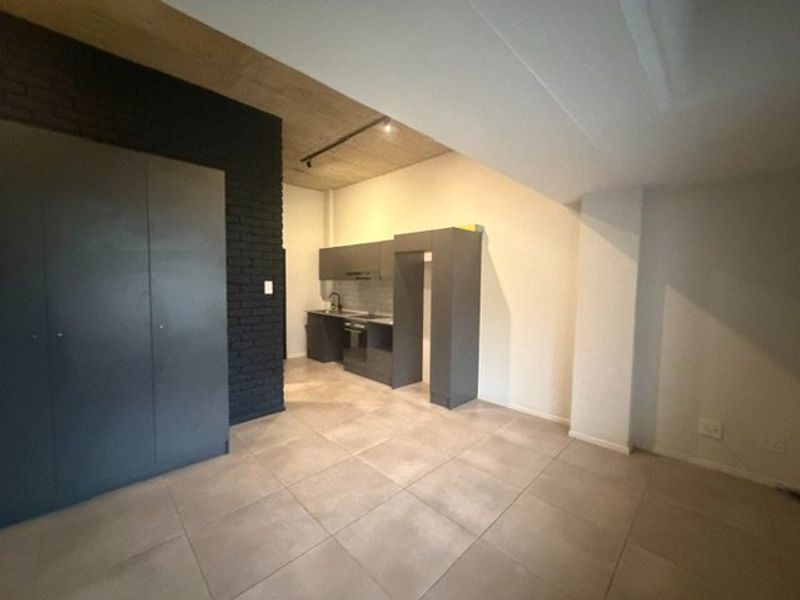 Modern open-plan living area featuring a sleek kitchen with built-in cabinetry, exposed concrete ceilings, and a striking dark brick feature wall.