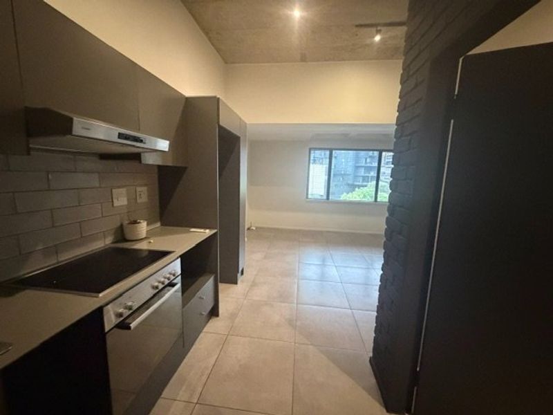 Modern open-plan kitchen and living area featuring sleek cabinetry, built-in appliances, tiled flooring, and an industrial-style exposed concrete ceiling and brick pillar.