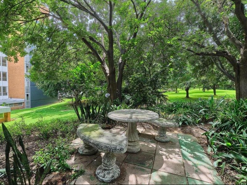 A tranquil garden area featuring mature trees, established greenery, and a paved seating area with stone furniture, adjacent to the apartment building.