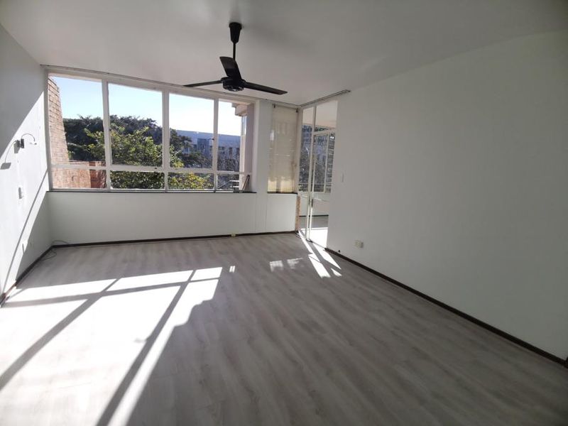 A bright, empty living room featuring light-toned laminate flooring, a ceiling fan, and large windows providing ample natural light.