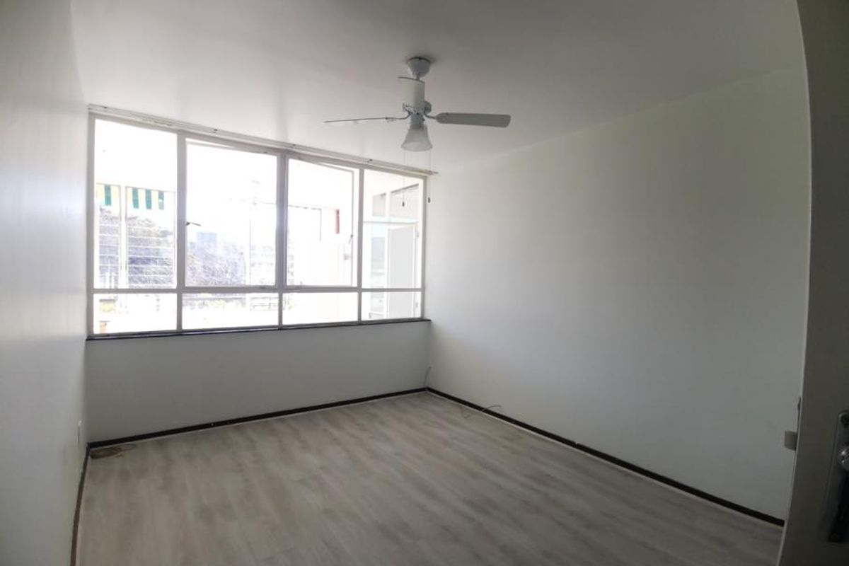 A bright, empty bedroom featuring light-toned laminate flooring, a ceiling fan, and large windows providing ample natural light.