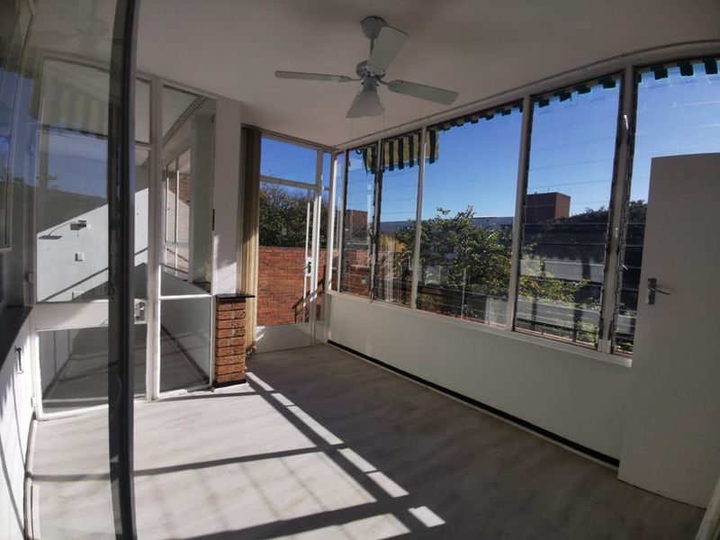 A bright, enclosed balcony or sunroom featuring large glass windows, laminate flooring, and a ceiling fan, offering a versatile space with natural light.