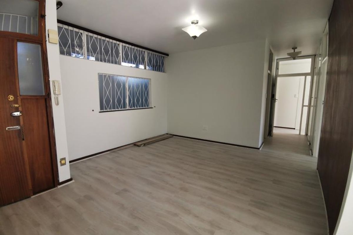 A bright, empty living room featuring modern laminate flooring, security bars on windows, and an integrated intercom system near the entrance.