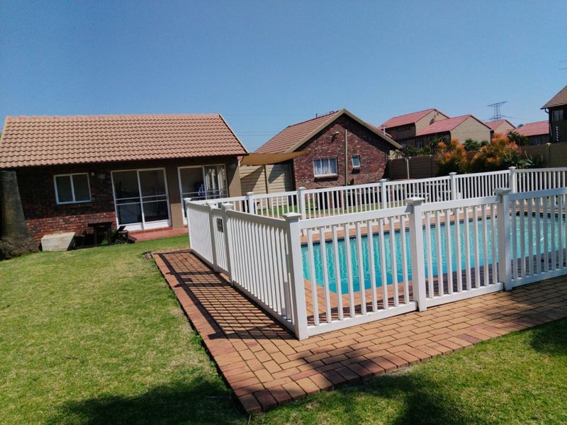 A well-maintained backyard featuring a swimming pool enclosed by a white picket fence, brick paving, and a lawn area adjacent to the main dwelling.