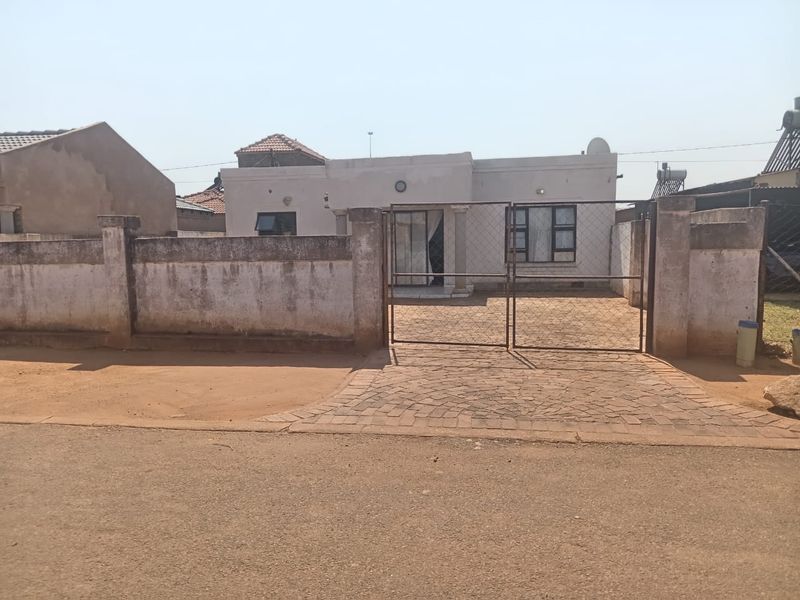 Front view of a single-story house featuring a paved driveway, metal gate, and perimeter wall with visible weathering and exterior finish wear.