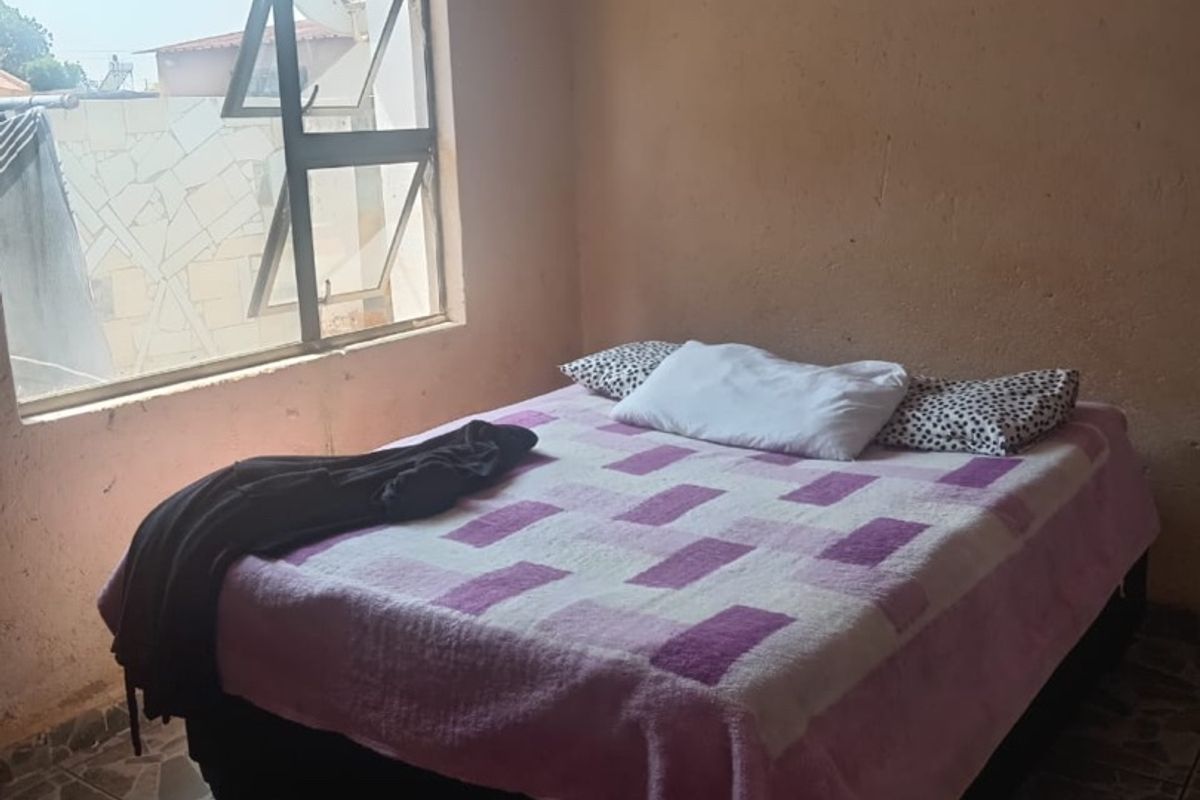 A bedroom featuring tiled flooring, an aluminium window frame, and a tongue and groove ceiling, currently showing signs of wear on the wall surfaces.
