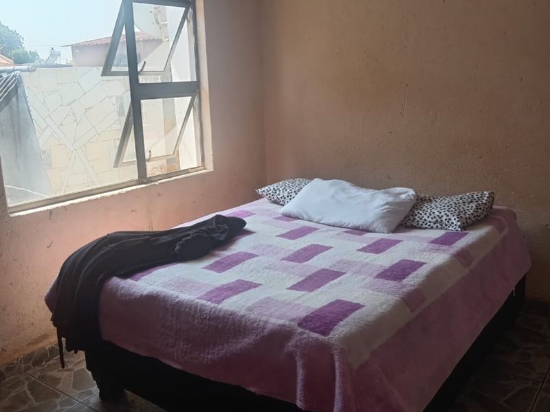 A bedroom featuring tiled flooring, an aluminium window frame, and a tongue and groove ceiling, currently showing signs of wear on the wall surfaces.