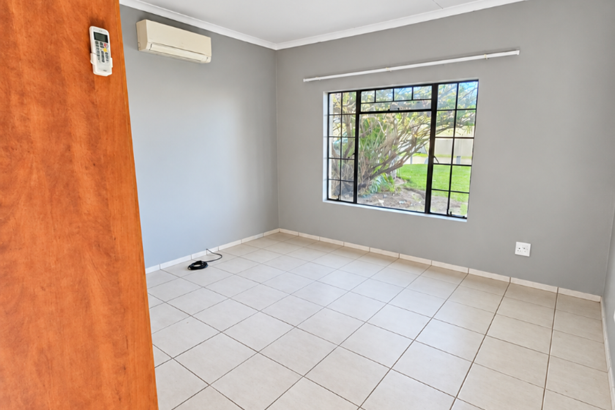 A bright, empty bedroom featuring neutral grey walls, tiled flooring, a split-system air conditioner, and a large window overlooking the garden.