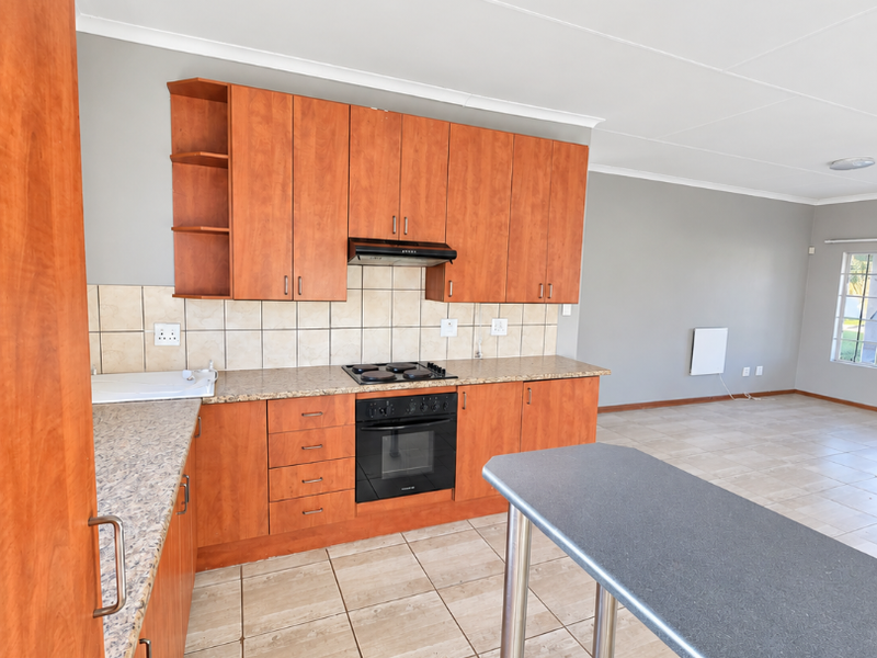 Open-plan kitchen featuring wood-finish cabinetry, laminate countertops, built-in oven and hob, and tiled flooring, flowing into an adjacent living area.