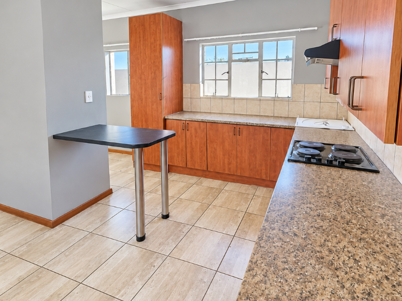A functional kitchen featuring wood-finish cabinetry, laminate countertops, an integrated electric hob with extractor fan, and neutral tiled flooring throughout.