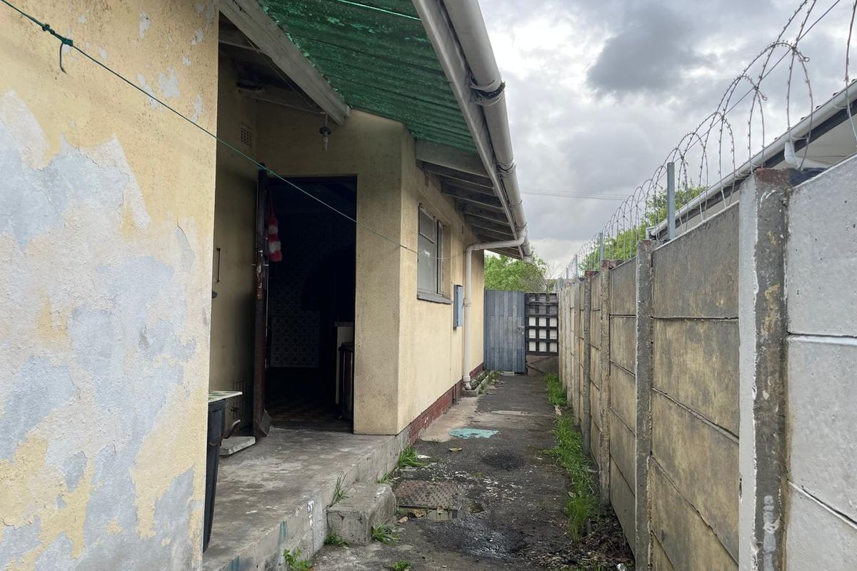 Rear exterior view of the property showing a concrete pathway, precast boundary wall with razor wire, and weathered exterior wall finishes.