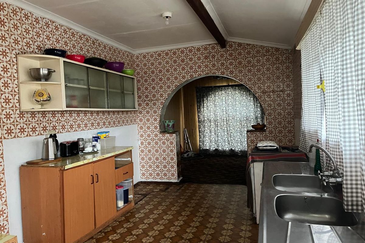 A dated kitchen featuring original patterned wall and floor tiles, built-in cabinetry, and a double stainless steel sink with an arched transition to the living area.