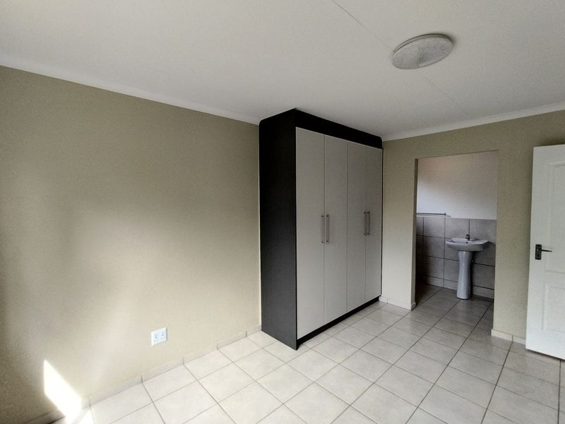 A clean, empty bedroom featuring tiled flooring, a modern built-in wardrobe, and an adjoining en-suite area with a pedestal basin.