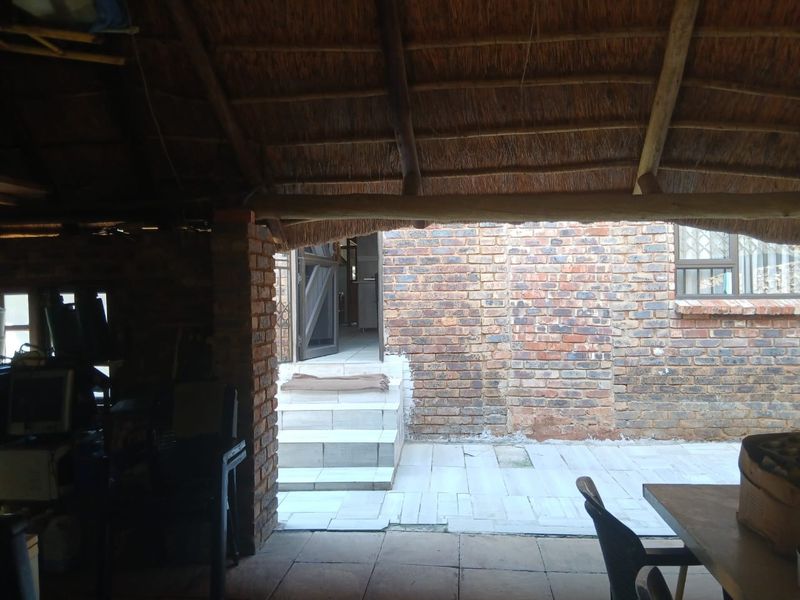 A covered outdoor patio area featuring a traditional thatch roof, exposed brick walls, and tiled flooring, providing a rustic transition to the main house entrance.