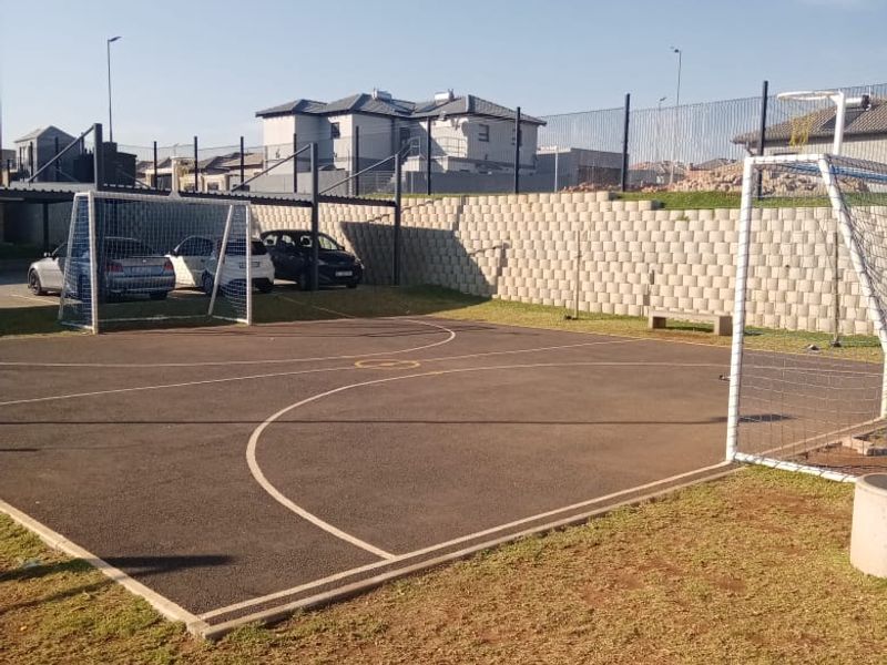 Outdoor sports facility featuring an asphalt court with soccer goals, enclosed by a retaining wall and perimeter fencing.