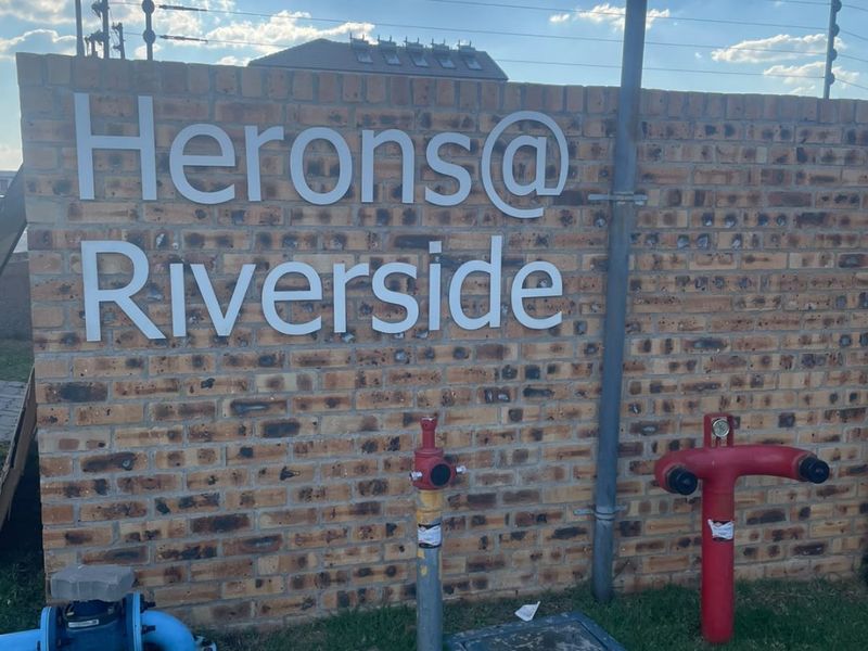 Exterior view of the Herons@Riverside complex entrance featuring a brick boundary wall, signage, and utility infrastructure.