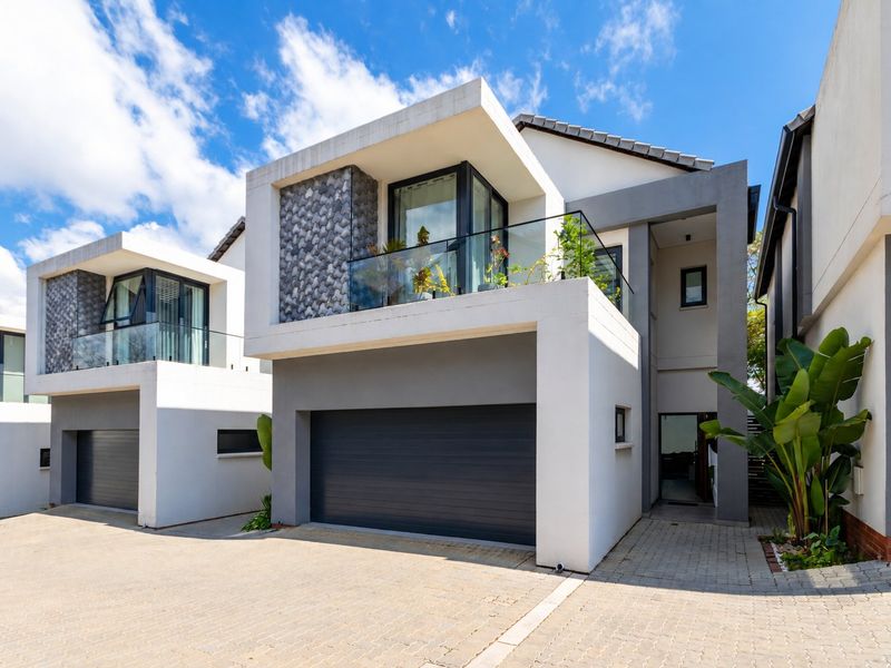 Modern double-storey townhouse facade featuring a sleek automated garage, glass-fronted balcony, and contemporary architectural finishes with paved driveway access.
