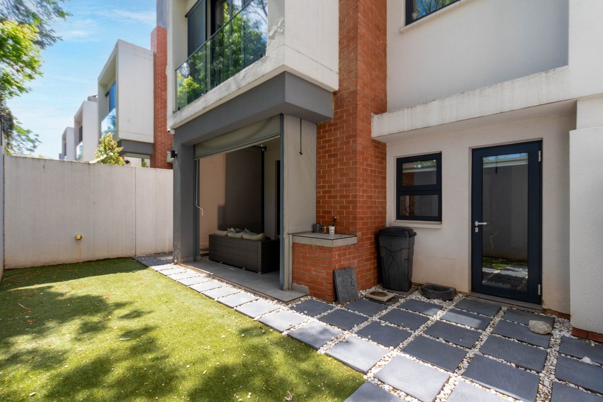 Rear exterior view featuring a paved patio area, artificial lawn, brick chimney detail, and access to the main dwelling via sliding and standard doors.