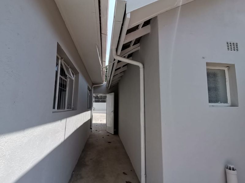 A narrow exterior side passage between two building sections featuring concrete flooring, white painted walls, and functional guttering systems.