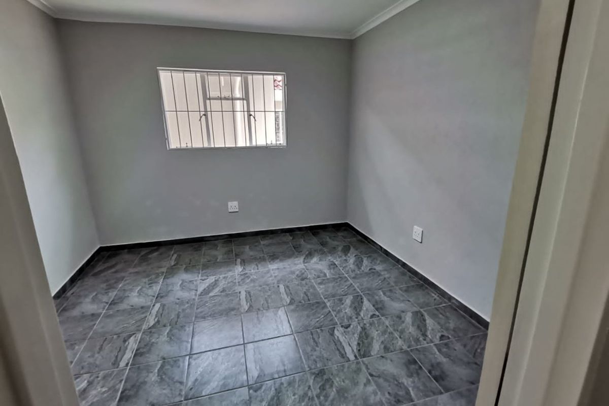 A clean, empty bedroom featuring modern grey tiled flooring, neutral walls, and a window fitted with security bars.