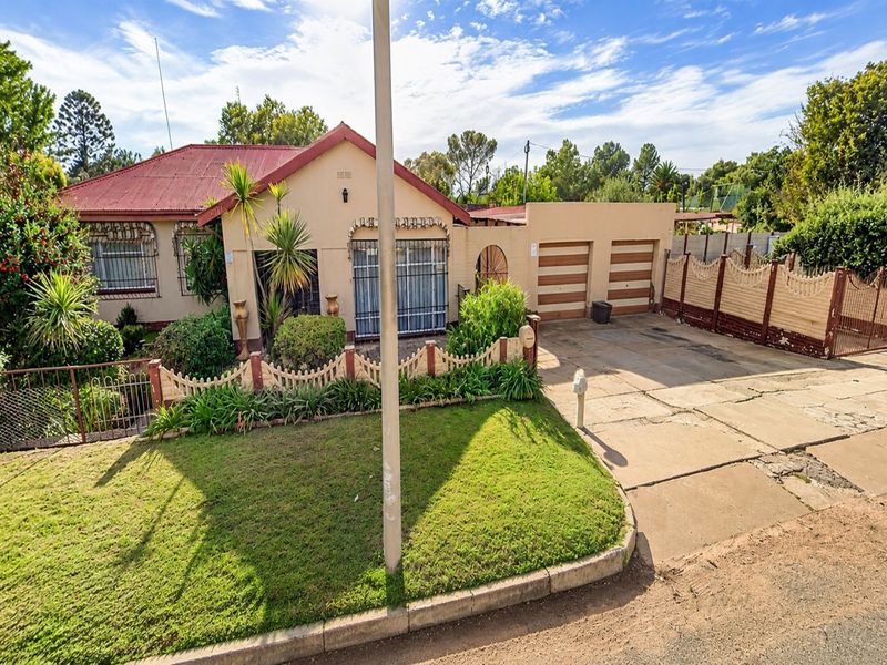 Front view of a single-story house featuring a corrugated metal roof, double garage, paved driveway, and established garden with security fencing.