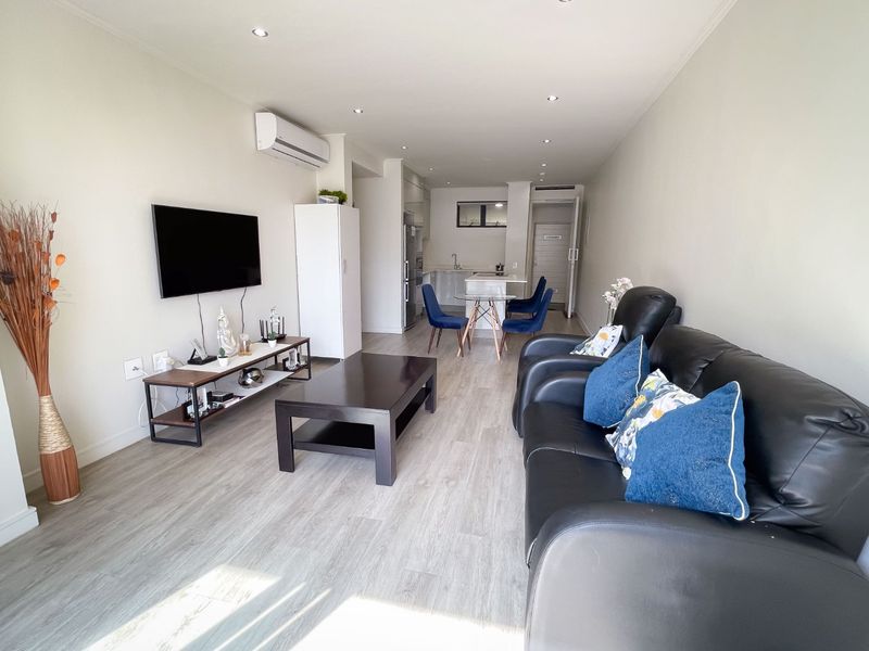 An open-plan living and kitchen area featuring modern laminate flooring, recessed downlighting, and a wall-mounted air conditioning unit.