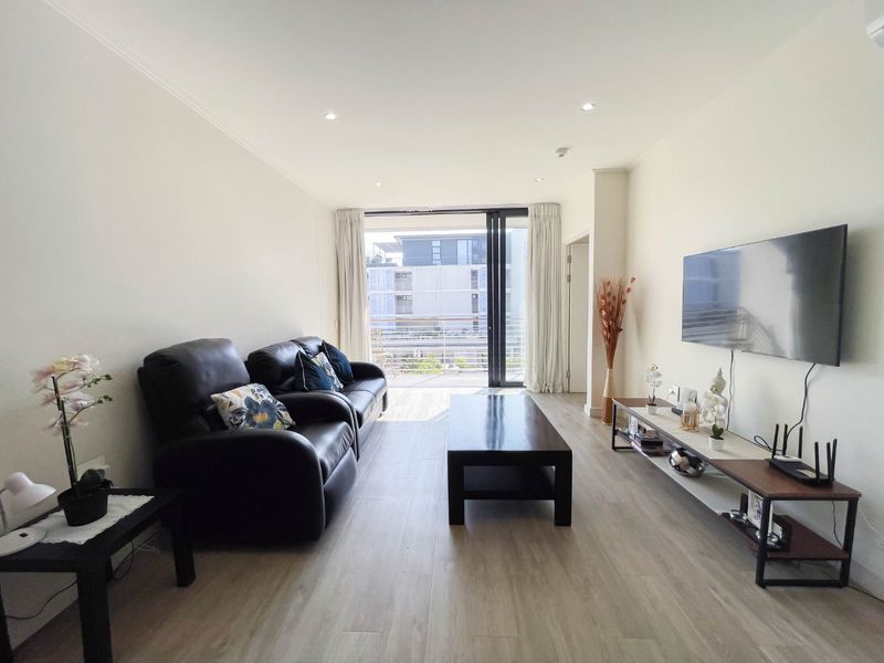 A bright living room featuring laminate flooring, recessed downlights, and a large sliding glass door providing access to the balcony.