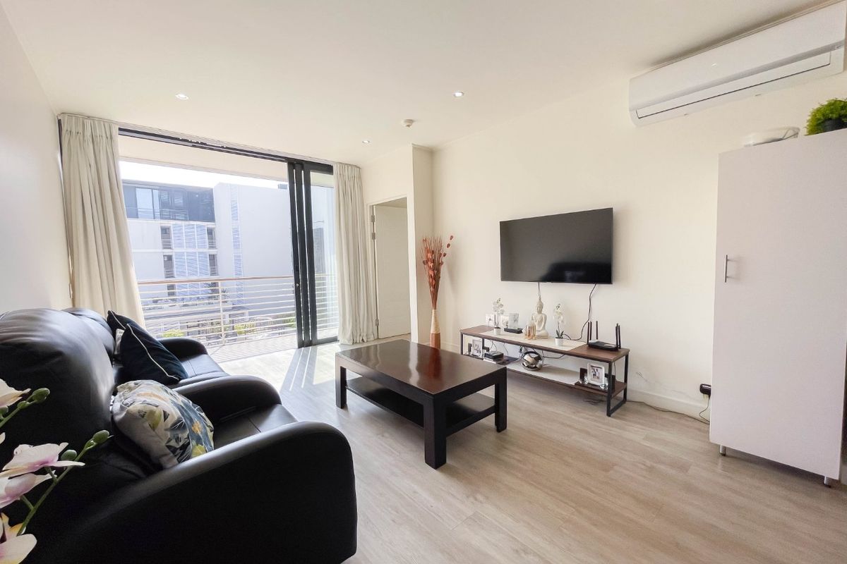 A bright living room featuring light laminate flooring, recessed downlighting, a wall-mounted air conditioning unit, and direct access to a balcony through sliding glass doors.