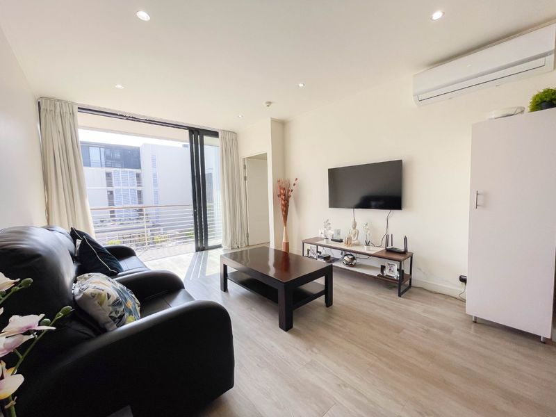 A bright living room featuring light laminate flooring, recessed downlighting, a wall-mounted air conditioning unit, and direct access to a balcony through sliding glass doors.