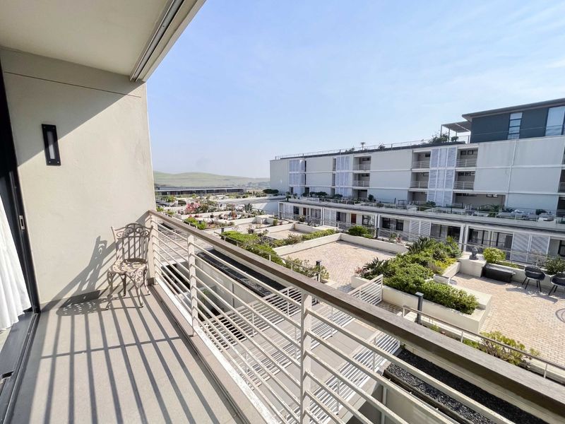 A spacious apartment balcony featuring a metal balustrade and views over the complex courtyard and surrounding landscape.