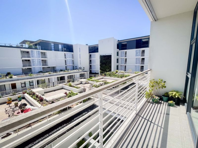 A spacious balcony with a metal balustrade, offering an elevated view over the apartment complex's landscaped courtyard and garden area.