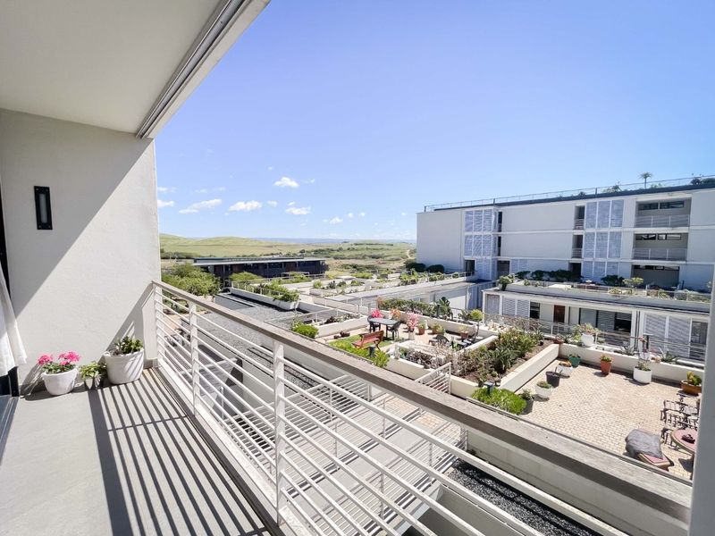 A clean, modern balcony with a metal balustrade, offering an elevated view over the apartment complex courtyard and surrounding landscape.
