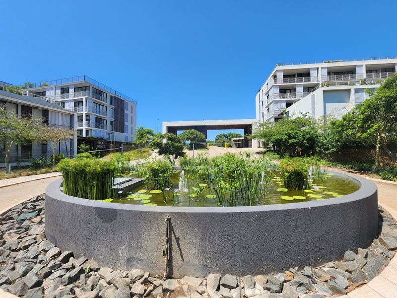 A well-maintained apartment complex entrance featuring a circular water feature, landscaped gardens, and modern multi-story residential buildings under a clear blue sky.