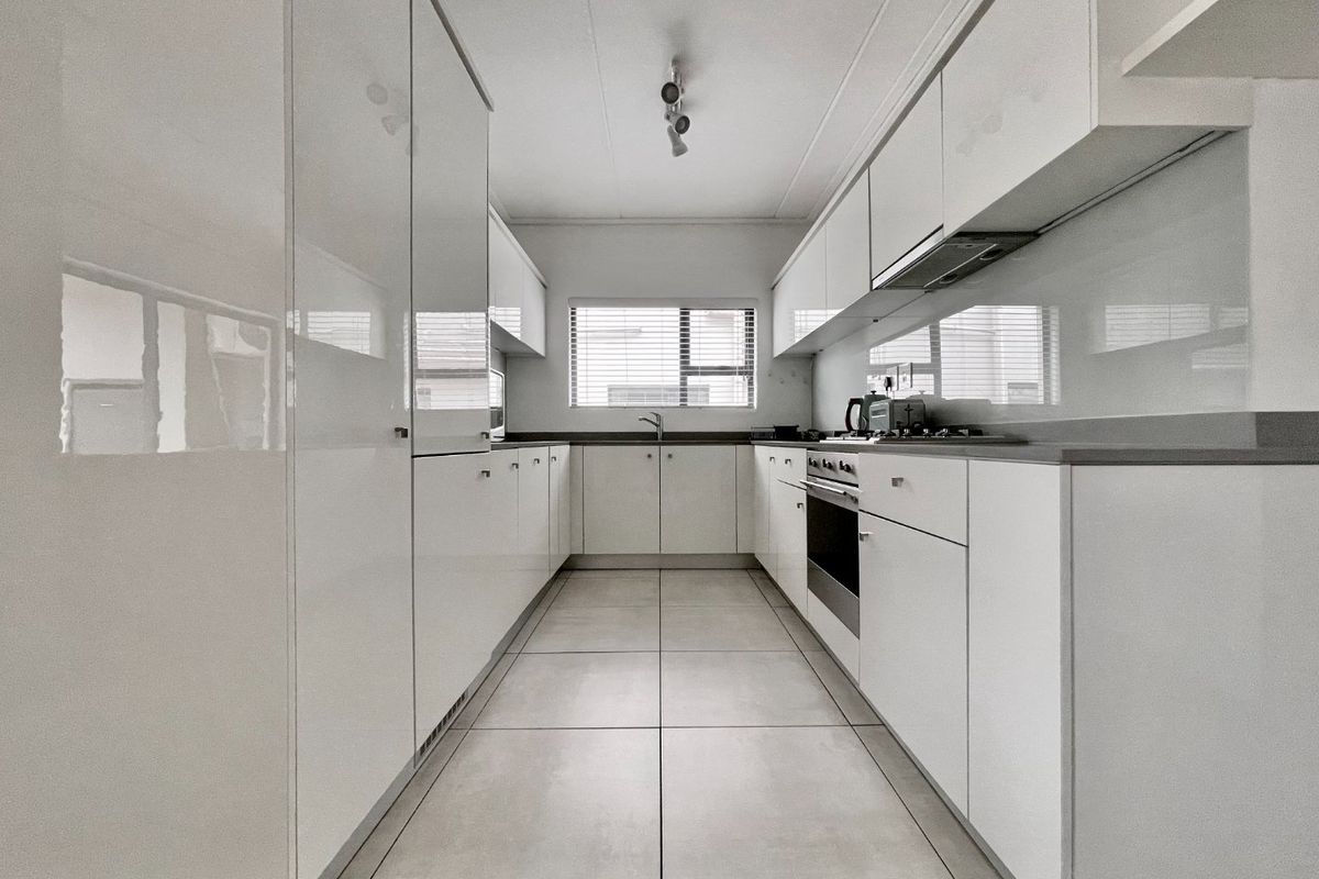 A modern, galley-style kitchen featuring sleek white cabinetry, stone countertops, integrated appliances, and large format floor tiles.