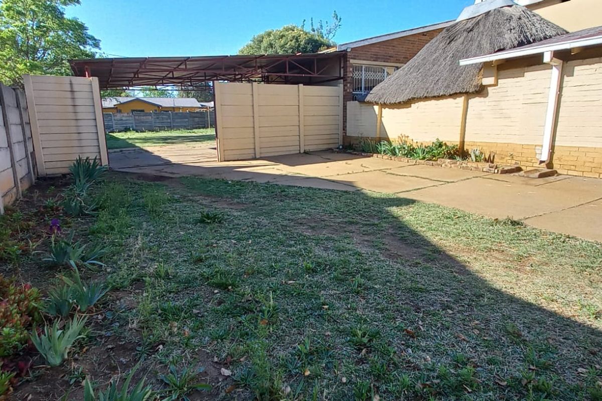 Rear exterior area featuring a concrete driveway, metal carport, and precast boundary wall, with a section of the main house showing a thatched roof.