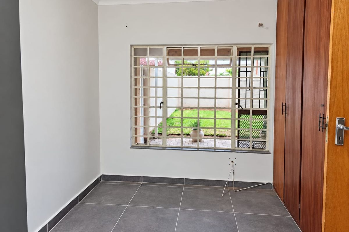 A clean, empty bedroom featuring tiled flooring, built-in wooden cupboards, and a window with security burglar bars overlooking the rear garden.