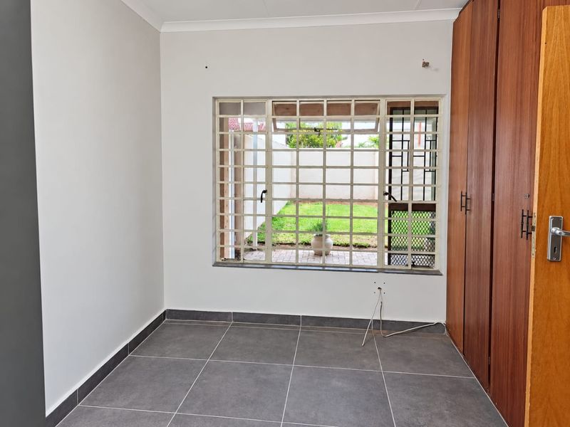 A clean, empty bedroom featuring tiled flooring, built-in wooden cupboards, and a window with security burglar bars overlooking the rear garden.