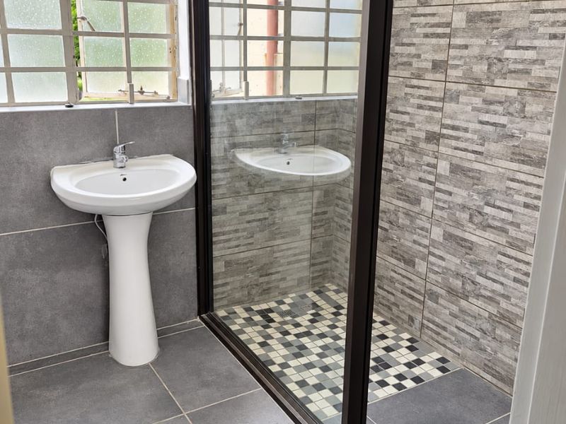 A modern bathroom featuring a pedestal basin, a glass-enclosed shower with mosaic floor tiles, and grey wall tiling throughout.
