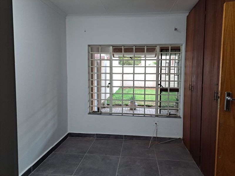 A vacant bedroom featuring tiled flooring, built-in wooden cupboards, and security bars on the window overlooking the rear garden.